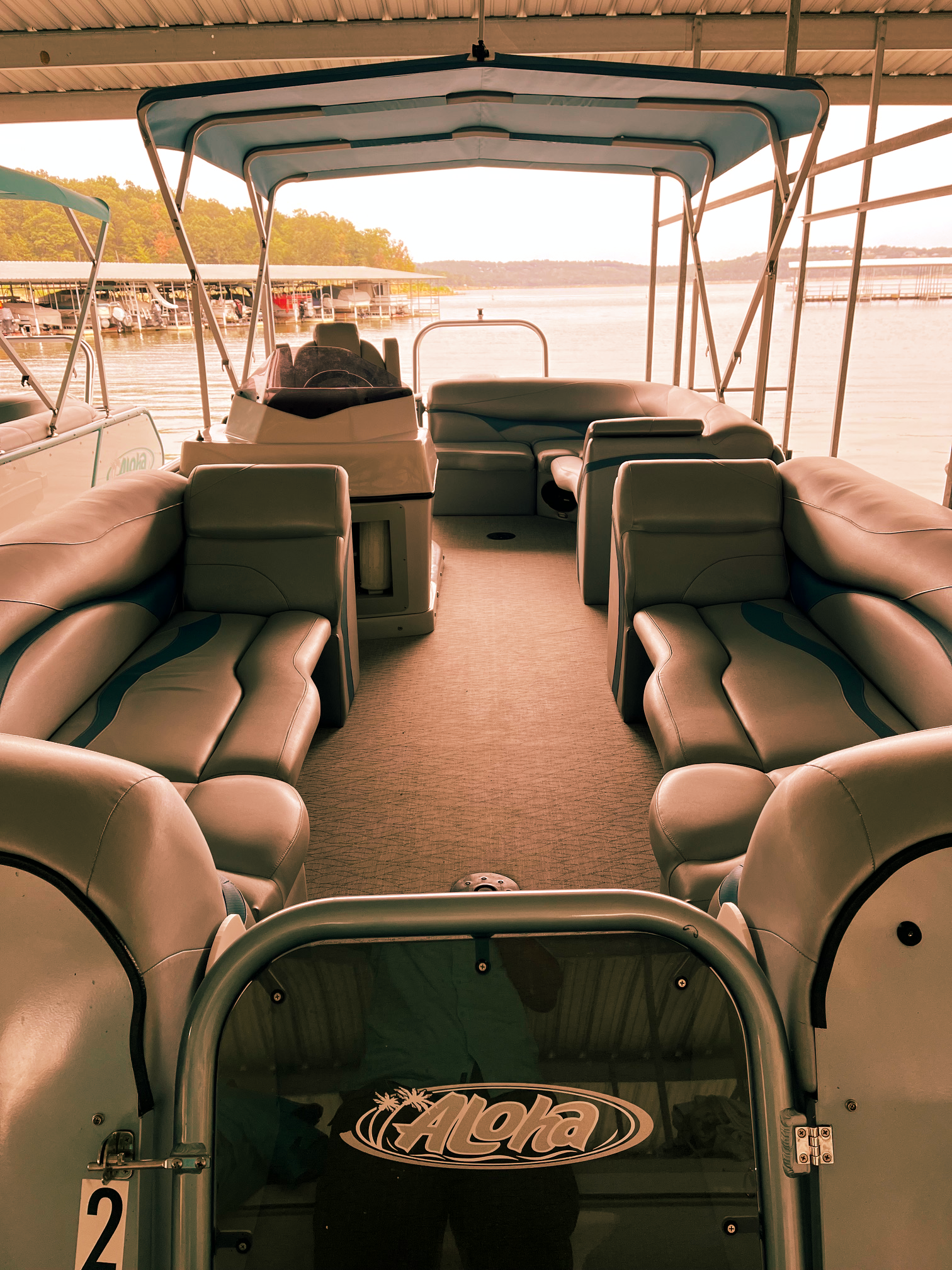 Interior view of a premium Aloha Tropical Series tritoon boat rental parked under a covered dock slip, showing plush grey and blue upholstery, parallel front lounge seating, center console helm, and an overhead bimini top looking out onto Norfork Lake.