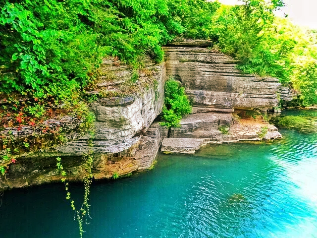 Clear, vibrant turquoise water of Norfork Lake running alongside towering, layered "chunky" limestone rock bluffs covered in lush green summer foliage.